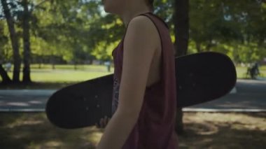 kaykaycı bir parkta elinde bir kaykay ile yürüyor