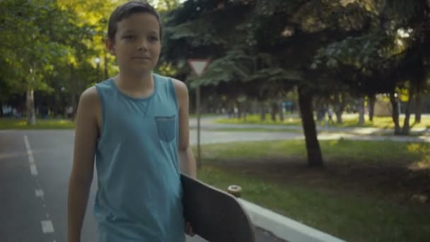 Skateboarder camina en un parque con un monopatín en las manos . — Vídeos de Stock