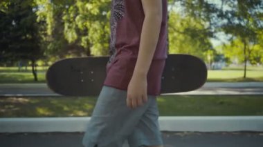 kaykaycı bir parkta elinde bir kaykay ile yürüyor.