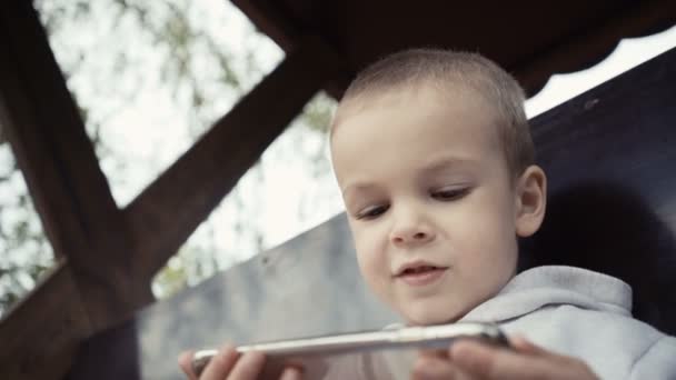 两年的男孩坐在公园的长凳上, 看着智能手机上的卡通片 — 图库视频影像