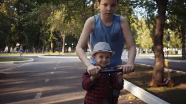 Dos hermanos montando un patinete scooter en un parque de verano . — Vídeos de Stock