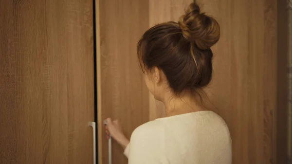 Woman choosing dress from cupboard in room. Attractive lady with brown hair looks to decide what to wear