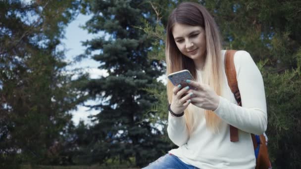 Filmati bella ragazza seduta vicino all'albero nel parco e utilizzando app sul suo smartphone . — Video Stock