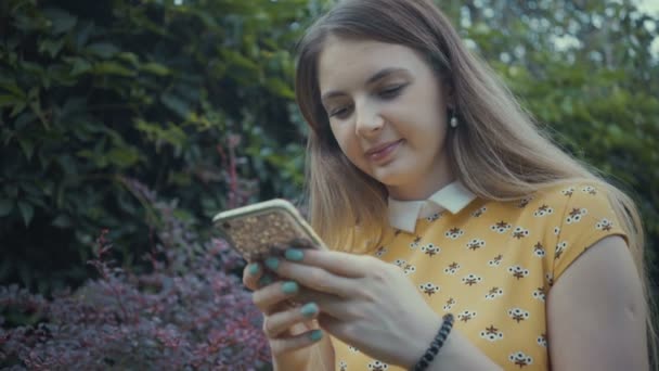 Filmación chica bonita utilizando la aplicación en su teléfono inteligente cerca del árbol en el parque . — Vídeo de stock