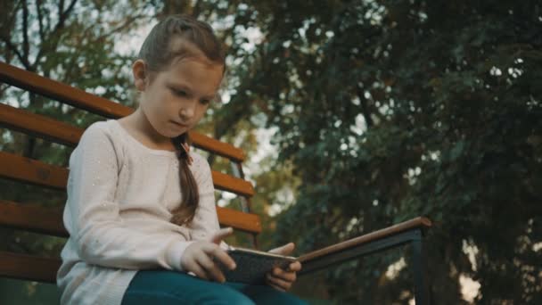 Écolière assise sur un banc en bois dans le parc et utiliser un smartphone . — Video