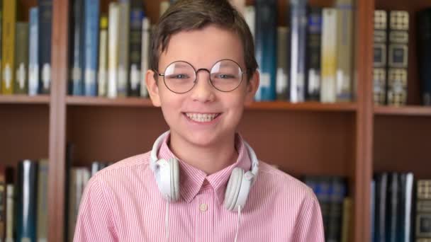 Портрет милого школьника, стоящего возле книжной полки в библиотеке — стоковое видео