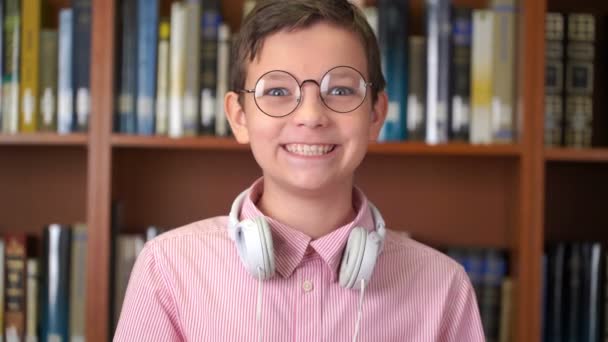 Портретний знімок милого школяра, що стоїть біля книжкової полиці в бібліотеці і робить великий палець вгору — стокове відео