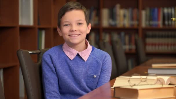 Porträtt skott av den söta skolpojke sitter nära bokhyllan i biblioteket — Stockvideo