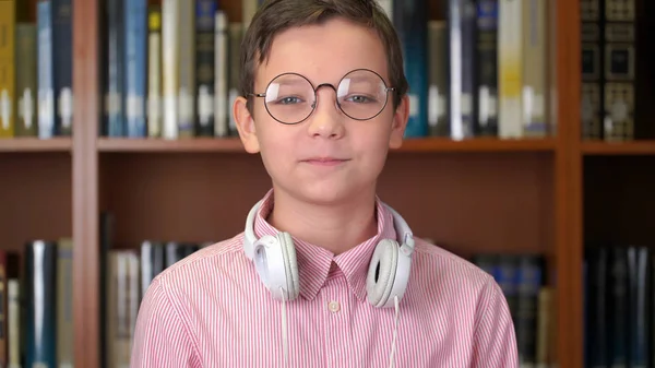 Retrato de la foto del colegial lindo de pie cerca de la estantería en la biblioteca . — Foto de Stock