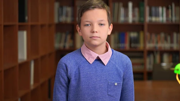 Portrait shot of the sad, unhappy schoolboy standing near the bookshelf in the library — Stock Photo, Image