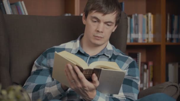 Портрет человека, читающего книгу возле книжной полки в библиотеке — стоковое видео