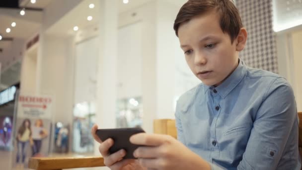 Lindo chico se sienta en el banco en el salón del topo y juega emocionalmente en un juego en su teléfono inteligente — Vídeo de stock