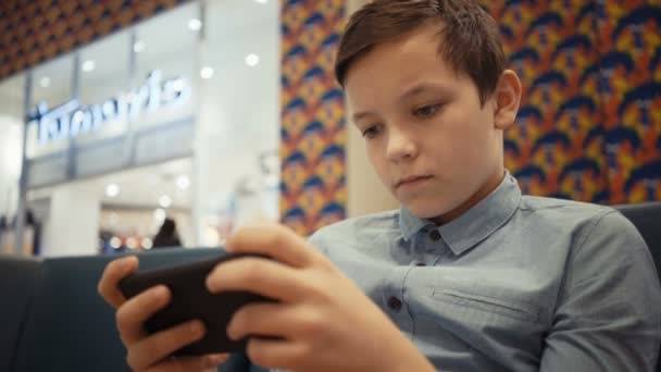 Lindo chico se sienta en el sofá en el salón del topo y juega emocionalmente en un juego en su teléfono inteligente — Vídeo de stock