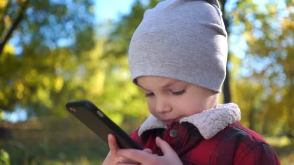 Jongen is speelspel op smartphone in herfst park. — Stockvideo