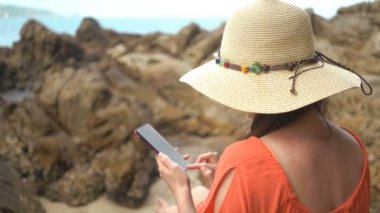 Genç güzel kadın sosyal ağlarda iletişim için telefon kullanarak ve denize yakın bir taş üzerinde oturan.
