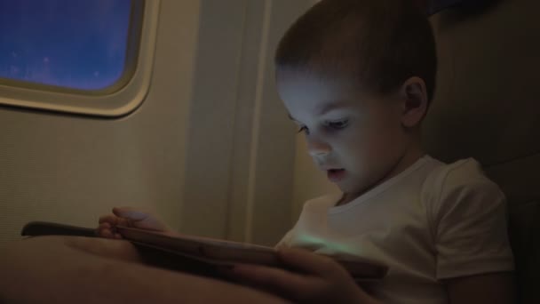 Милый мальчик путешествует в самолете и играет с планшетом — стоковое видео