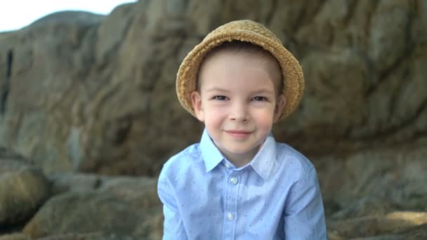 Cute boy sits on the shore against the backdrop of large stones. — Stock Video