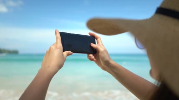 Νεαρή ταξιδιώτης γυναίκα στο καπέλο λήψη φωτογραφιών στο smartphone στην παραλία του ωκεανού — Αρχείο Βίντεο