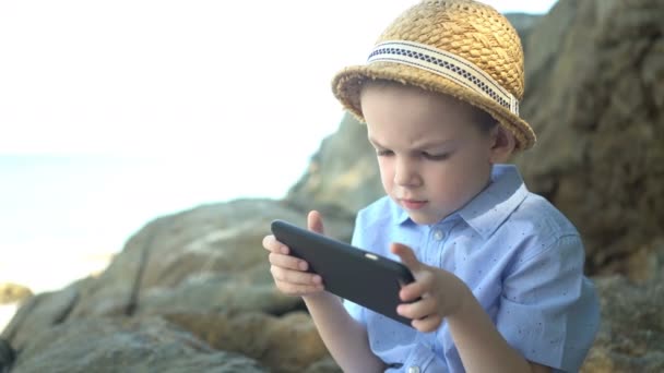 Lindo chico sentado en una piedra, usando un teléfono inteligente y mirando la aplicación del teléfono cerca del mar — Vídeo de stock