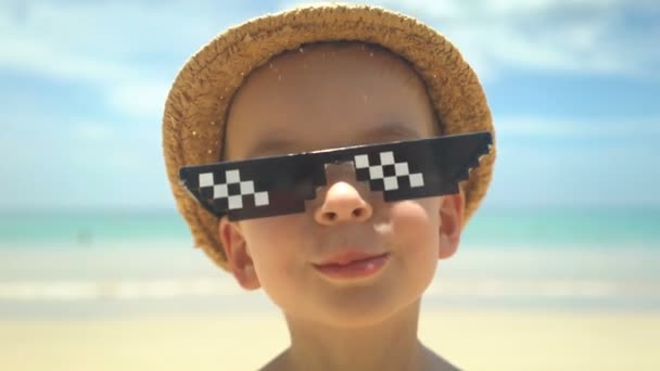Portrait de mignon petit garçon en chapeau de paille avec des lunettes de soleil debout sur la plage d'été . — Video