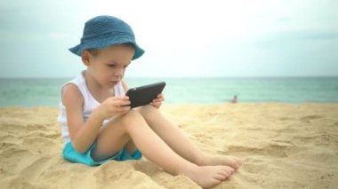 Kumlu plajda oturan ve akıllı telefon uygulamasını kullanan sevimli çocuk