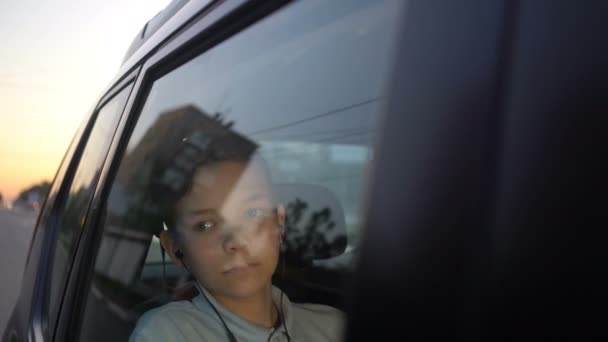 Menino adolescente com fones de ouvido ouvindo a música no banco de trás do carro e olhando para longe — Vídeo de Stock