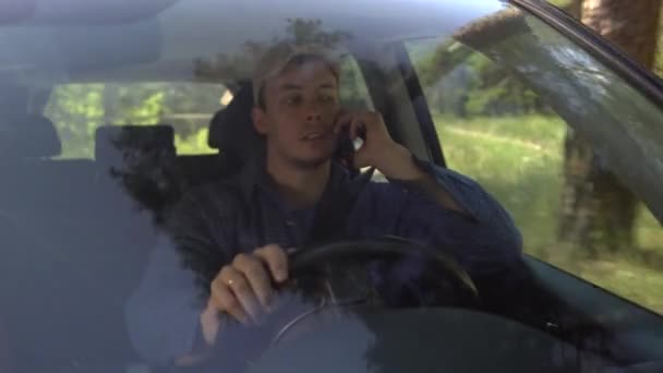 Young man looking around driving car along forest side on summer day and talking on the phone — Stock Video