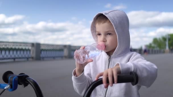 かわいい3-4歳の男の子は、最初のランニングバランスバイクに乗って水を飲むことを学びます — ストック動画