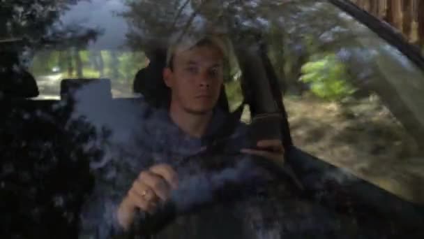 Hombre joven mirando alrededor de coche de conducción a lo largo del lado del bosque en el día de verano y el uso de teléfono inteligente — Vídeo de stock