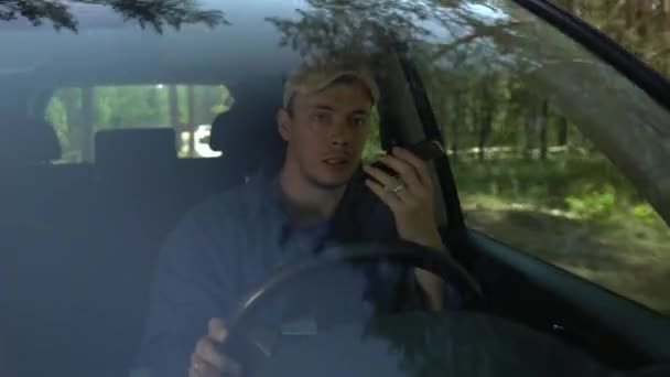 Молодой человек смотрит вокруг вождения автомобиля вдоль лесной стороне в летний день и говорить по телефону — стоковое видео