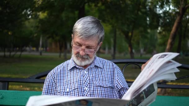 Sênior em camisa xadrez senta-se em um banco no parque e lê um jornal — Vídeo de Stock