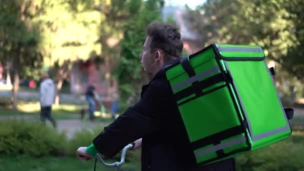 Repartidor con mochila verde pasea en bicicleta por la ciudad con comida a domicilio — Vídeos de Stock