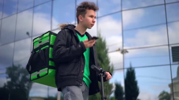 Homem de entrega com bolsa verde e uma scooter está procurando o endereço dos clientes no telefone — Vídeo de Stock