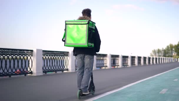Repartidor con mochila verde monta un scooter por la ciudad con comida a domicilio — Vídeos de Stock