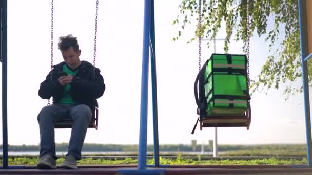 Курьер с зеленым багажом ждет заказа на доставку на качелях — стоковое видео