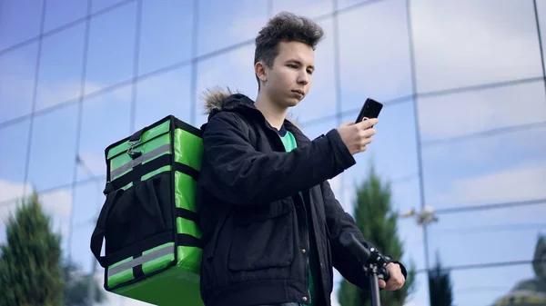 delivery man with green backpack and a scooter is looking for the clients address in the phone