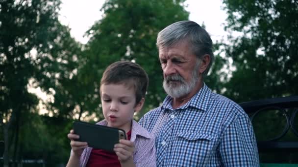 Ο παππούς και ο εγγονός του κάθονται σε ένα παγκάκι στο πάρκο και παίζουν σε ένα smartphone. — Αρχείο Βίντεο