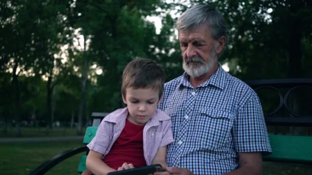 Starszy dziadek i wnuk siedzą na ławce w parku i grają na smartfonie — Wideo stockowe