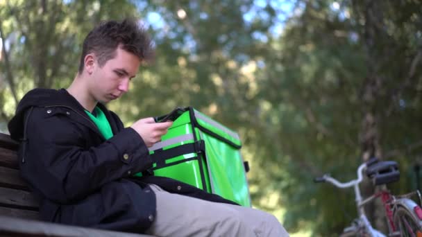 Fattorino con zaino verde e una bicicletta sta cercando l'indirizzo del cliente al telefono — Video Stock