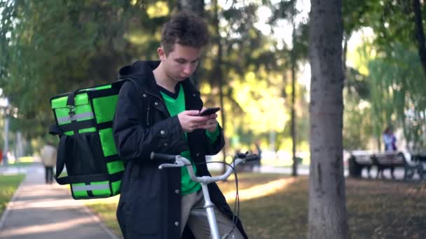 Homem de entrega com mochila verde e uma bicicleta está procurando o endereço dos clientes no telefone — Vídeo de Stock