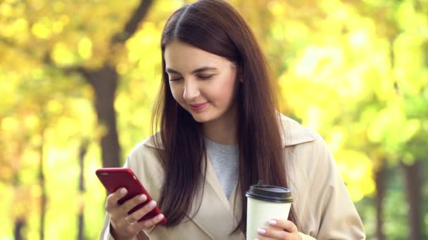 屋外の女性は、彼女のコーヒーと携帯電話で公園でコートやトレンチを身に着けている — ストック動画