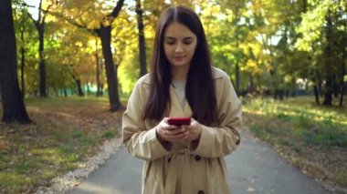 Sonbahar parkında yürüyen akıllı telefon kullanarak palto veya siper giyen genç kadın