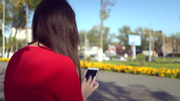 Atractiva mujer de negocios en vestido rojo caminando por la ciudad y utilizando la aplicación en su teléfono inteligente — Vídeo de stock