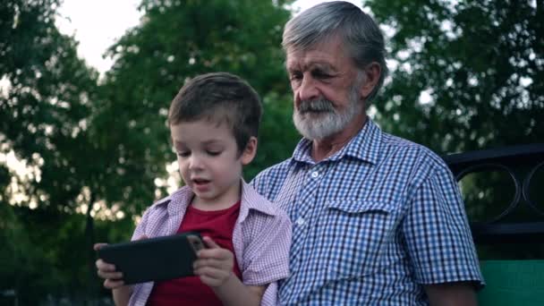 Avô e neto sênior estão sentados em um banco no parque e jogando em um smartphone — Vídeo de Stock