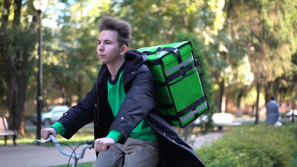 Livreur avec sac vert monte un vélo à travers la ville avec livraison de nourriture — Photo