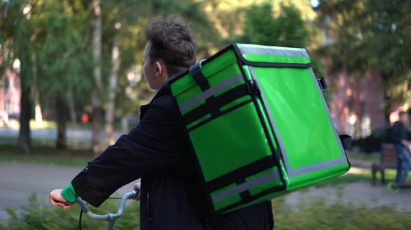 Repartidor con mochila verde pasea en bicicleta por la ciudad con comida a domicilio — Foto de Stock