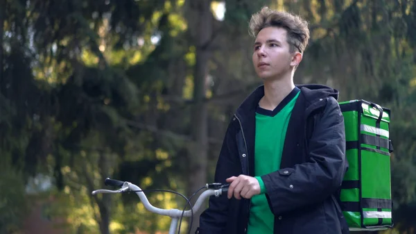 Repartidor con mochila verde pasea en bicicleta por la ciudad con comida a domicilio — Foto de Stock
