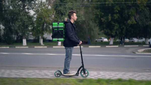 Homem de entrega com mochila verde monta uma scooter através da cidade com entrega de alimentos — Fotografia de Stock