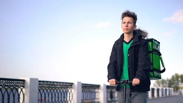 Repartidor con mochila verde monta un scooter a través de la ciudad con entrega de alimentos — Foto de Stock