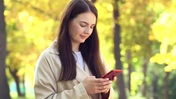 Mladá žena s kabátem nebo zákopu používající smartphone stojící na podzimním parku — Stock video